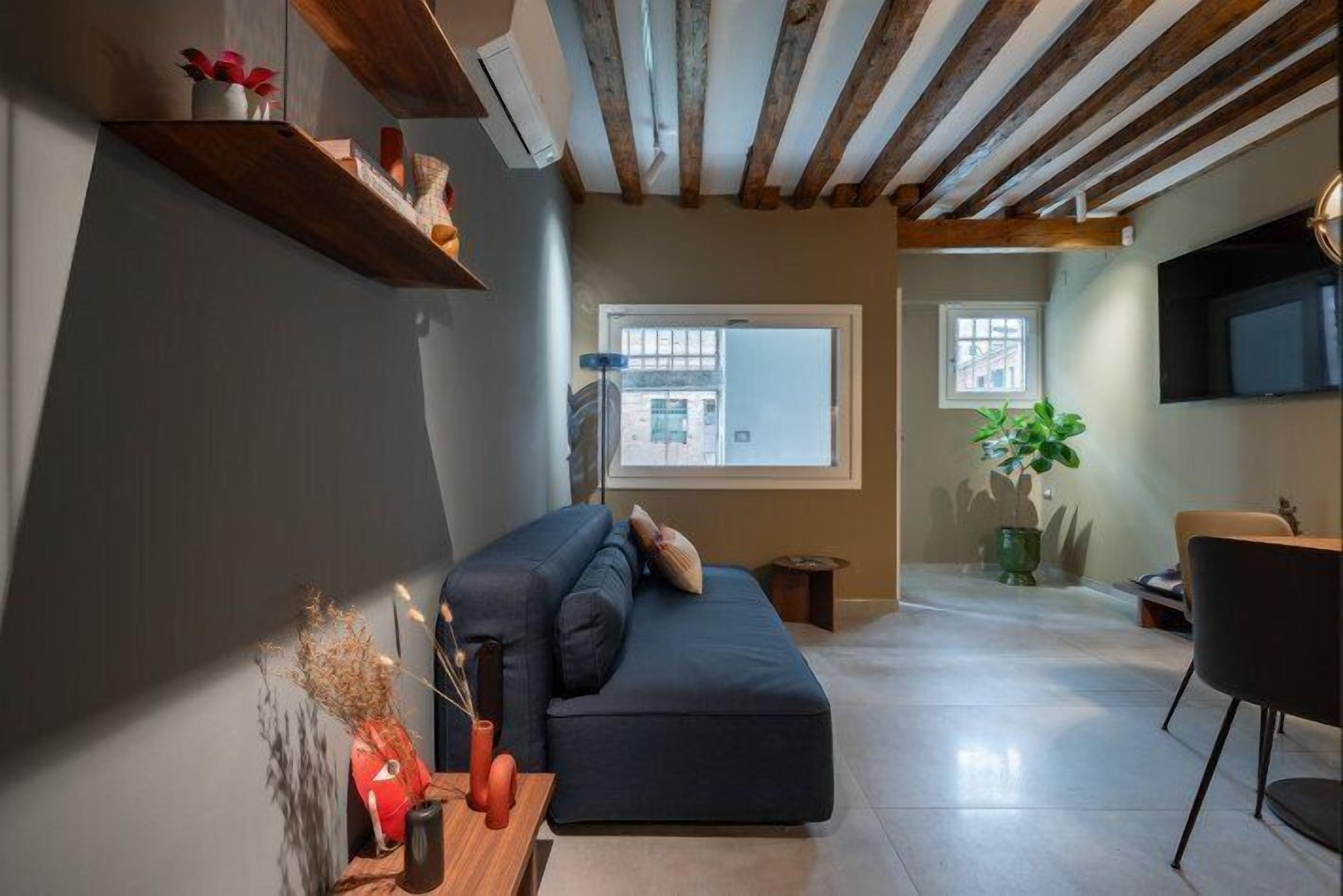 A modern living room with a dark blue sofa, wooden beams on the ceiling, and a large window providing natural light.