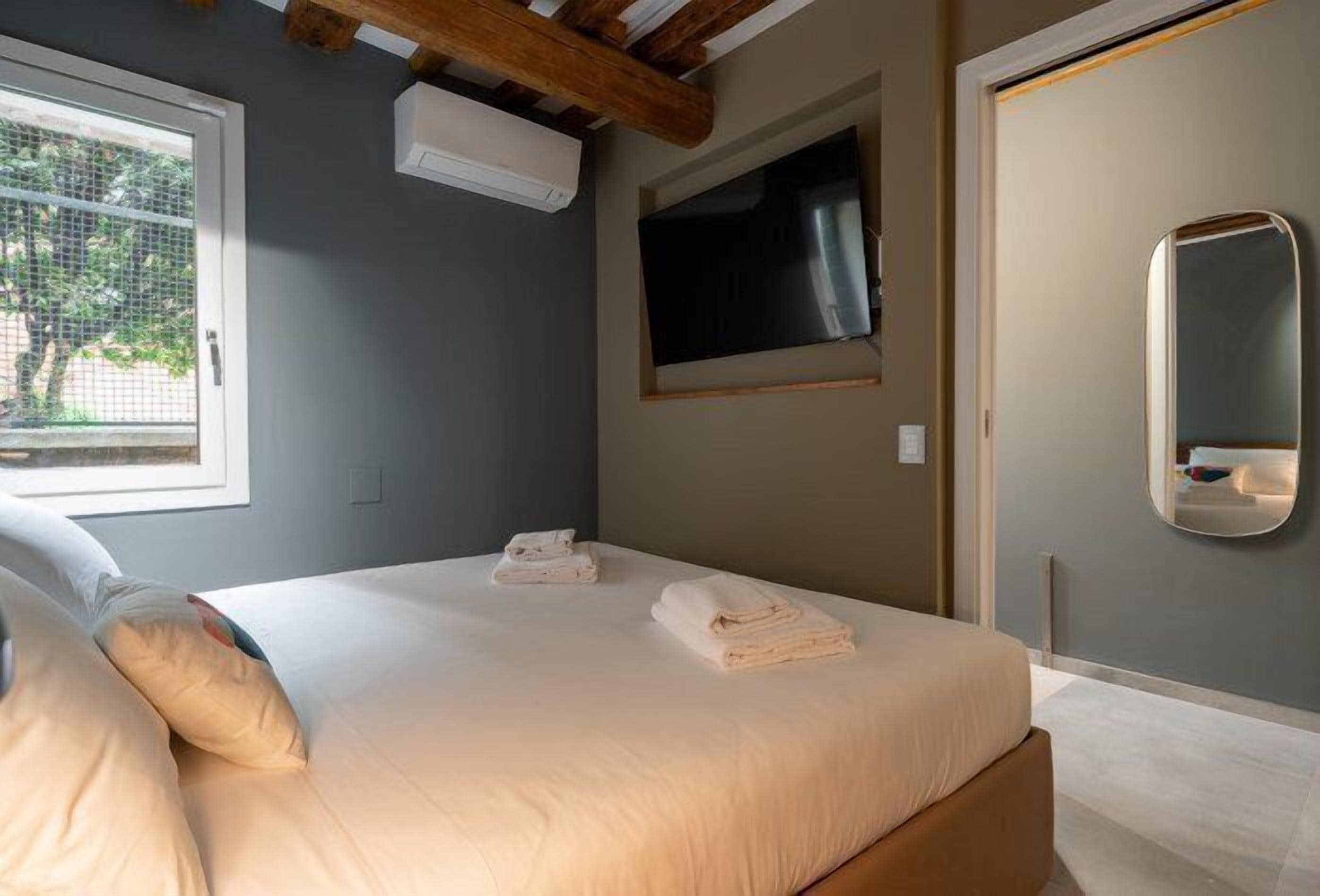 A neatly made bed with folded towels, a window with a view of trees, and a mounted TV.