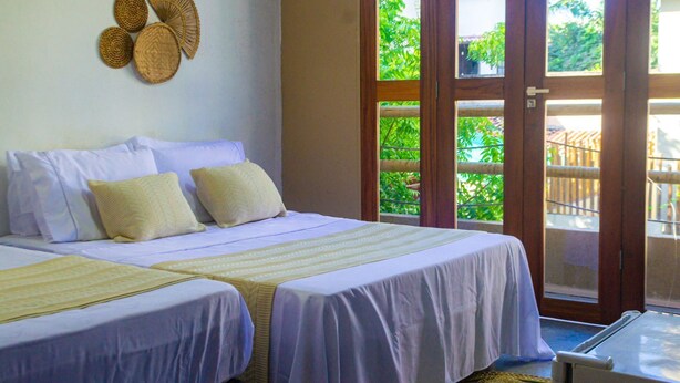 A bedroom with a bed, pillows, a bedside table, and a view of greenery outside. - Casa Mar Self Jeri (Jijoca de Jericoacoara)
