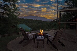 A fire pit with two Adirondack chairs and a table, surrounded by a gravel area with a view of a mountain range under a cloudy sky. - Romantic, cute and comfy rustic cabin with amazing view! (Sevierville)
