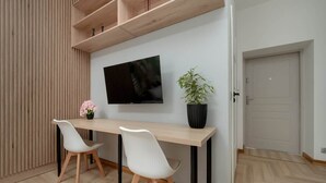 Una habitación moderna con suelo de madera, un televisor blanco montado en la pared, un escritorio de madera con dos sillas blancas y una planta en maceta.