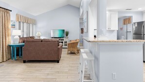 A modern living room with a brown sofa, a TV, and a dining area with a kitchen in the background. - Lakefront 2BR Beach Villa! GOLF CART, Near Beach, Pool, Tram, Screened Porch. (Miramar Beach)