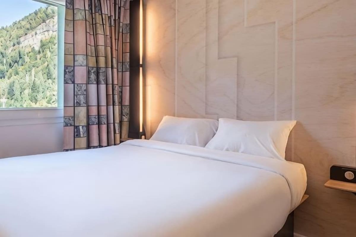 A hotel room with a bed, a nightstand, a wall-mounted clock, and a window with curtains.