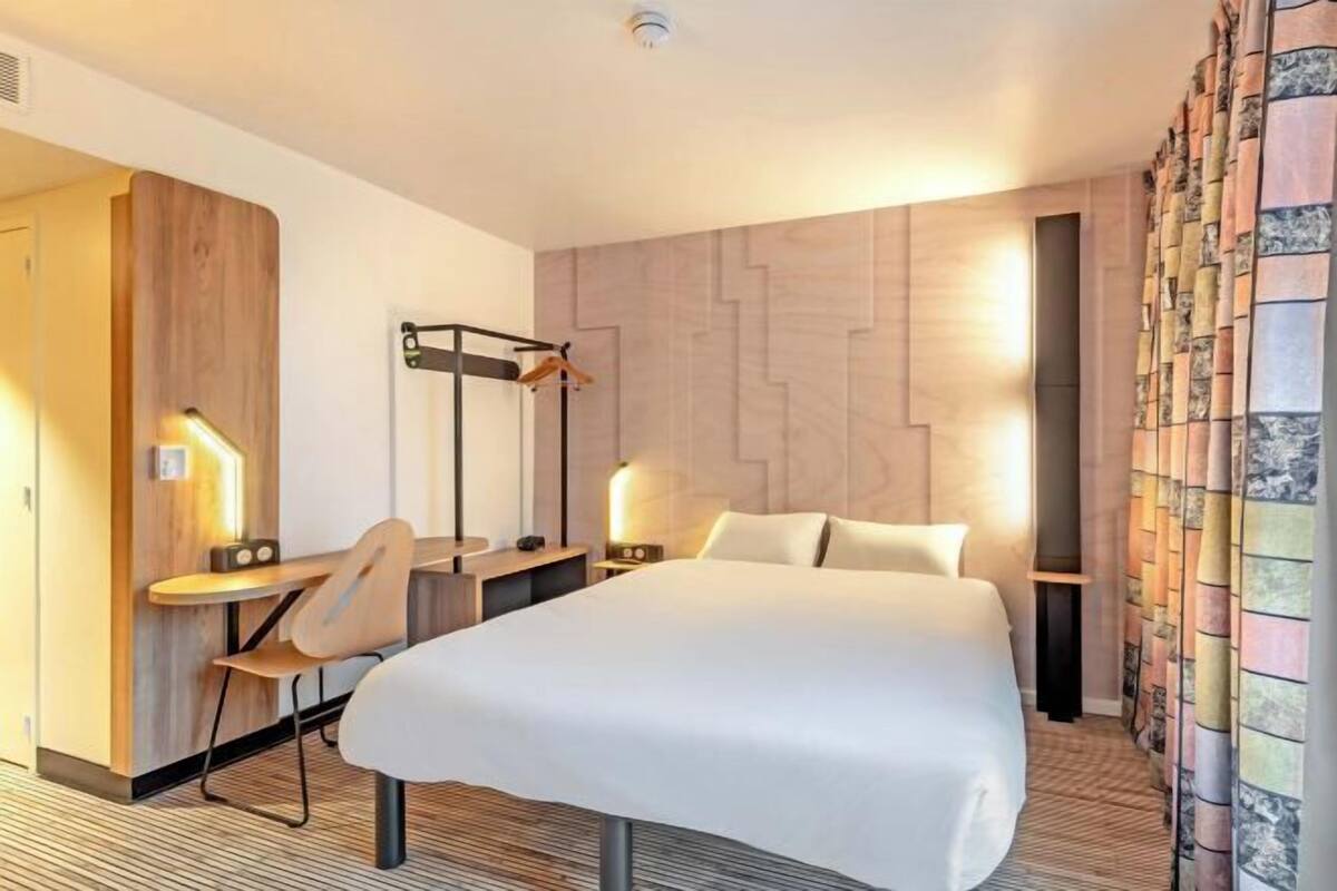 Hotellihuone, jossa on sänky, yöpöydät, työpöytä, tuoli, lamppu ja verho.