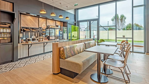 A modern cafe with a long counter, a seating area with a bench, and large windows offering a view of the outdoors. - B&B Hotel Brest Porte De Gouesnou (Gouesnou)
