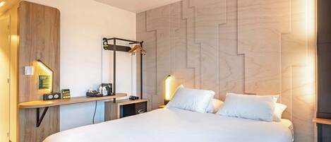 A hotel room with a large bed, a wooden headboard, a desk with a lamp, and a wall-mounted coat rack.