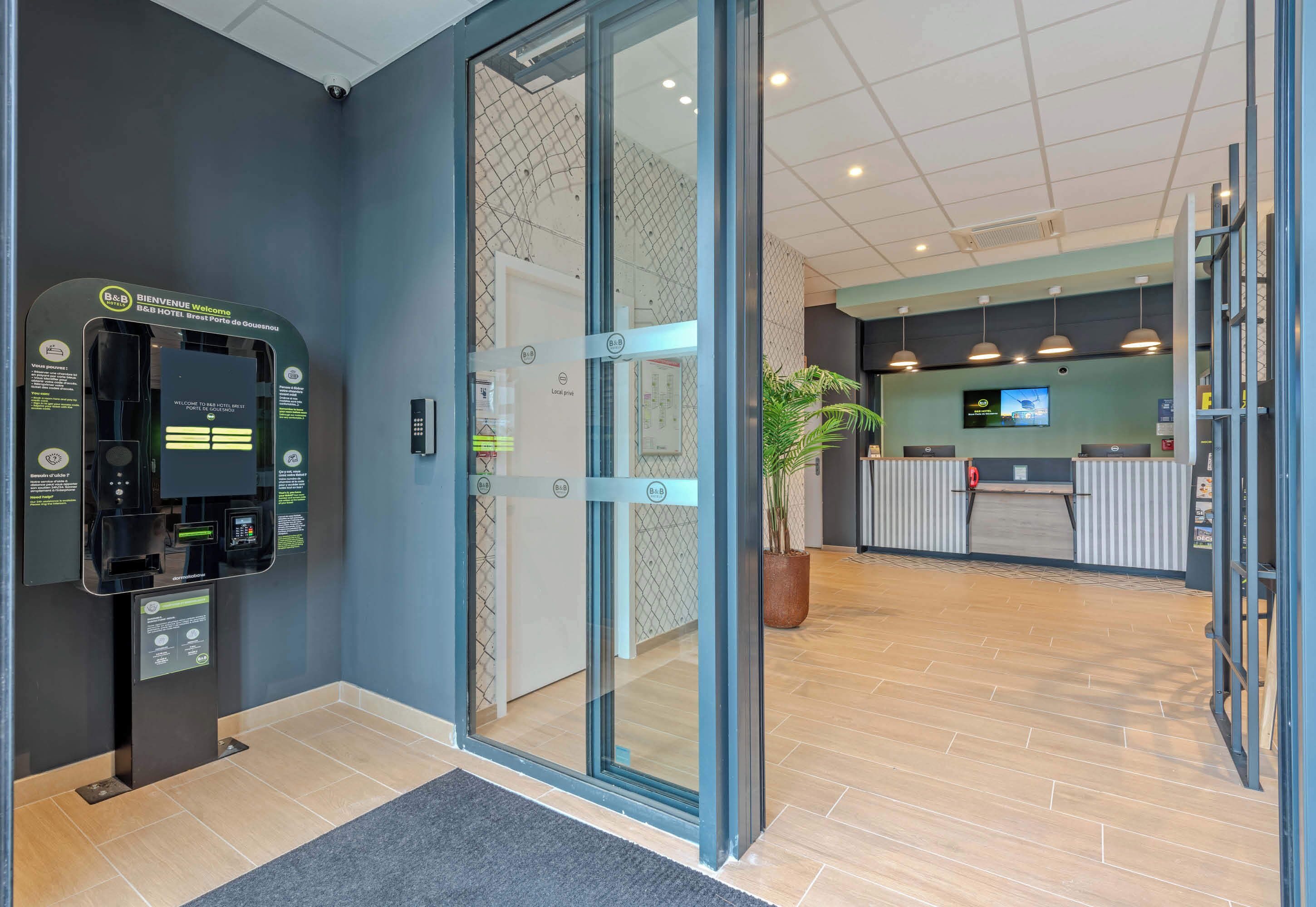 A hotel reception area with a self-service kiosk, a reception desk, and a potted plant.
