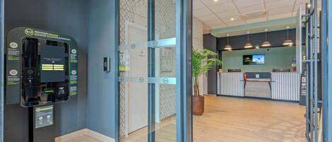 Un espace d’accueil d’hôtel comprenant un kiosque en libre-service, un comptoir d’accueil et une plante en pot.