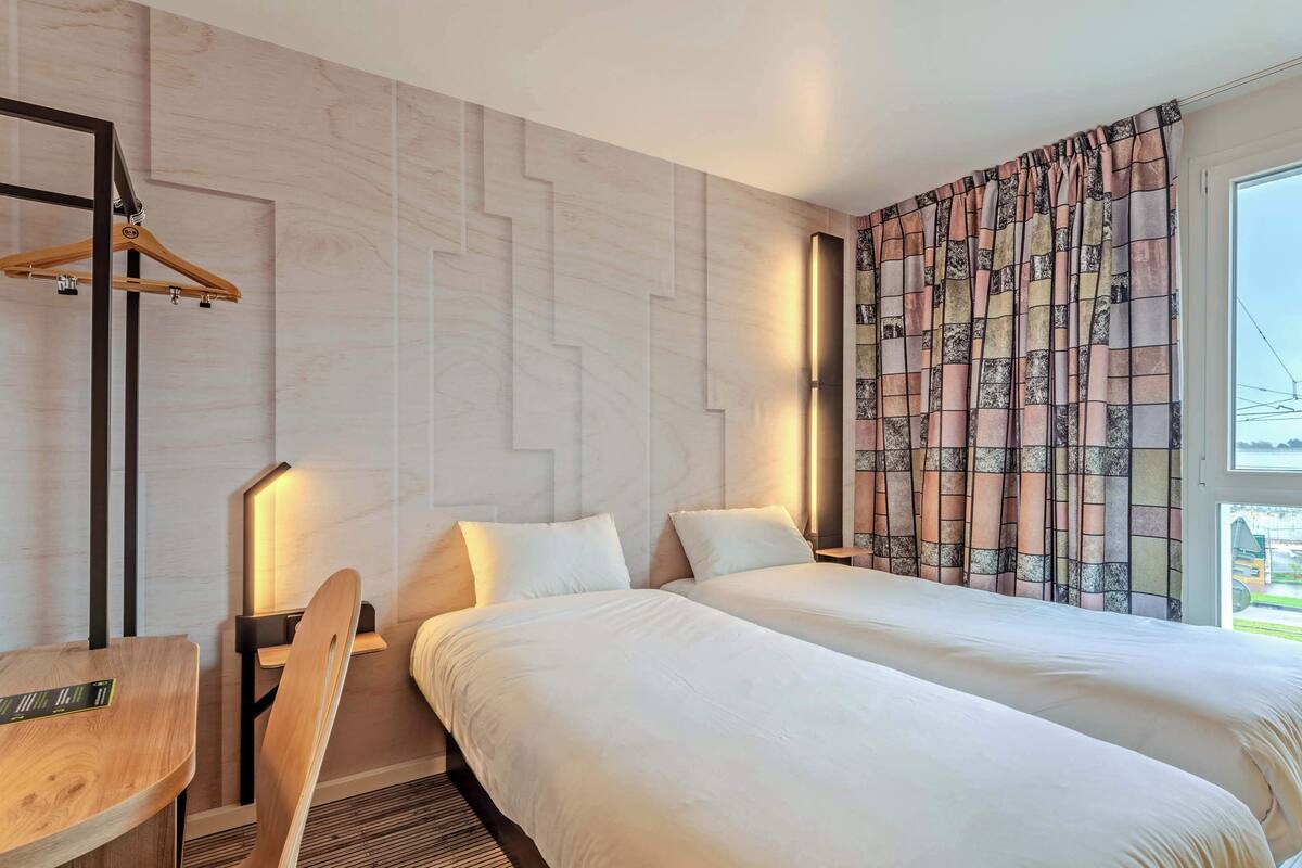 Hotellihuone, jossa on kaksi sänkyä, puinen työpöytä, tuoli, seinälle ripustettava naulakko ja suuri ikkuna, jossa on verhot.