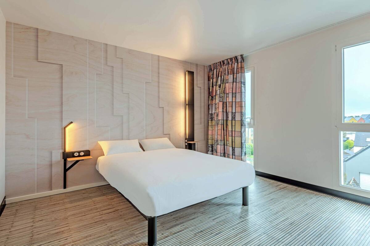 Moderni hotellihuone, jossa on suuri sänky, yöpöytä lampun kanssa, seinään kiinnitetty valaisin ja ikkuna, jossa on verhot.