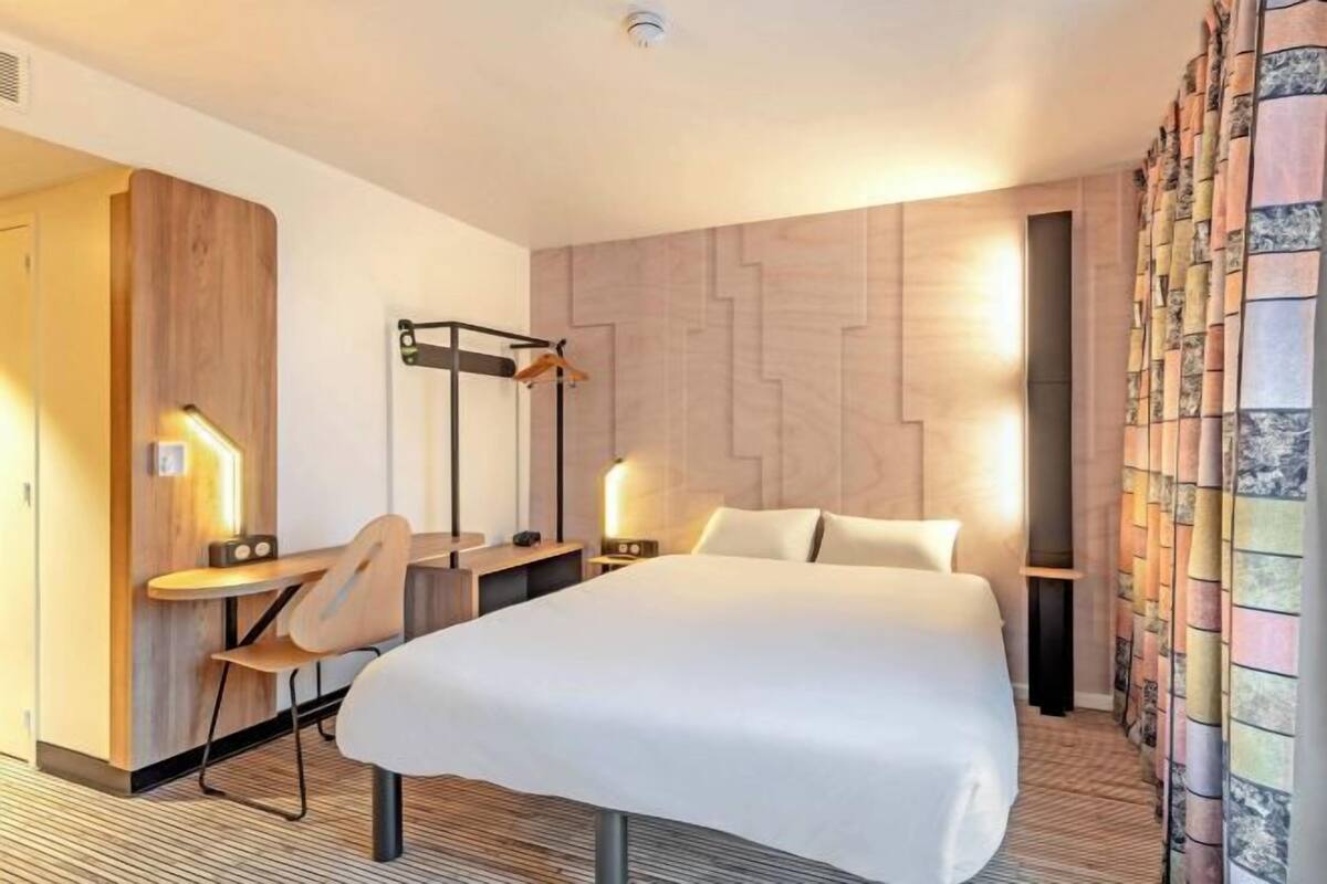 Hotellihuone, jossa on sänky, yöpöydät, työpöytä, tuoli ja verho.