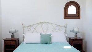 Una cama con cabecera blanca, dos mesitas de noche con lámparas, un almohón verde y una pequeña ventana sobre la cama.