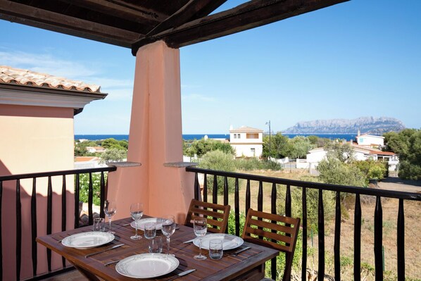 A wooden table set for dining with plates, glasses, and cutlery, overlooking a coastal landscape with buildings and a mountain in the distance. - Casa Le Sirene by Wonderful Italy (Olbia)