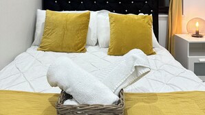 A neatly made bed with white and yellow bedding, a wicker basket with a white towel, and a bedside table with a lit candle.