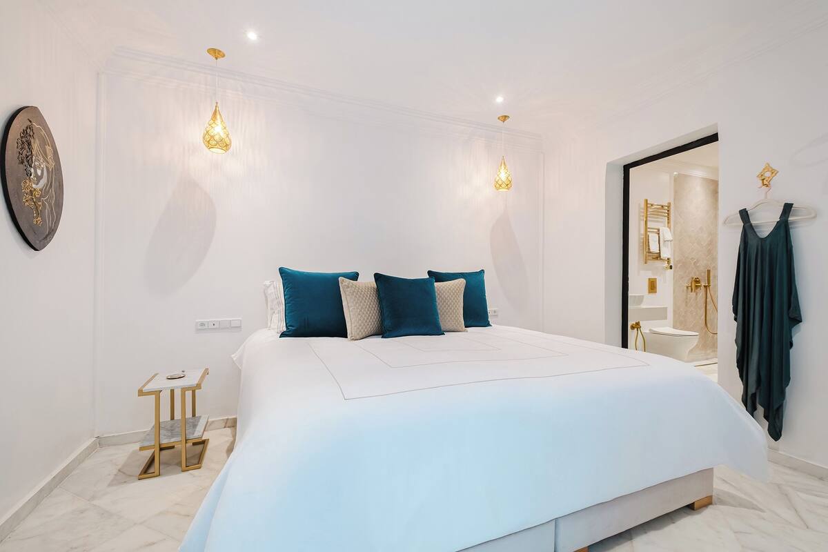 A modern bedroom with a large bed, two pendant lights, a small side table, and a hanging garment.