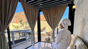 A person sitting on a white sofa with gold embroidery, in a room with large windows, a glass table with candles, and a view of a desert landscape. - Free People Riad and Spa (Marrakech)