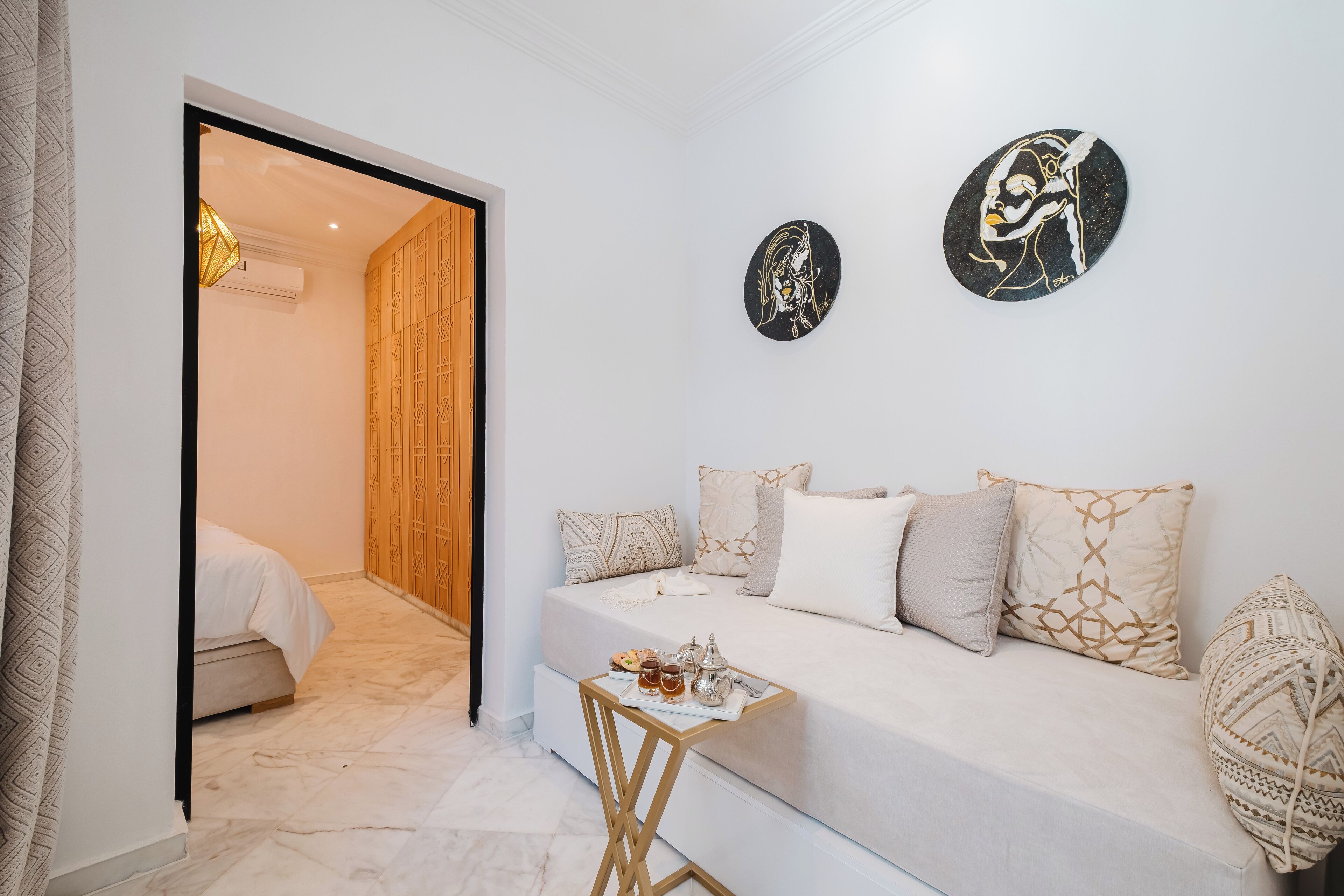 Un dormitorio moderno con un sofá blanco, almohadones estampados, una mesita de noche con bebidas y dos obras de arte en la pared.