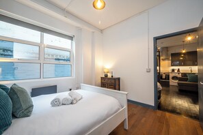 A bedroom with a large window, a bed with pillows, a bedside table with a lamp, and a view of a kitchen and laundry area. - Stylish 1 Bedroom Flat - Sleeps 4 (London)