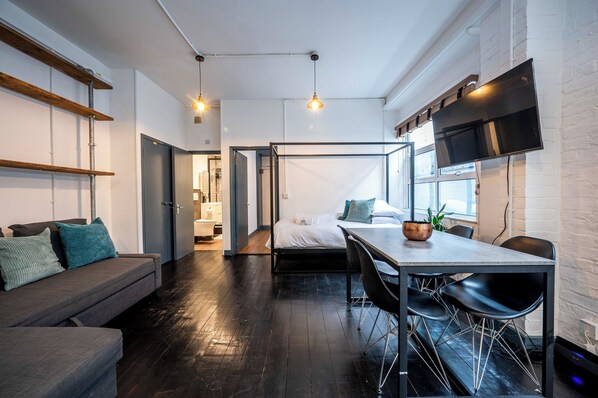 A modern, compact room with a bed, a sofa, a dining table with chairs, and a TV mounted on the wall. - Stylish 1 Bedroom Flat - Sleeps 4 (London)