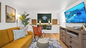 Un salon moderne avec un canapé jaune, un pouf rond, un téléviseur et un mur orné d’une affiche sur laquelle on peut lire : « Seules les bonnes ondes sont les bienvenues ».