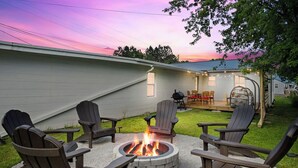 Un espace extérieur accueillant avec des fauteuils Adirondack disposés autour d’un foyer, une terrasse avec des chaises et une maison en arrière-plan.