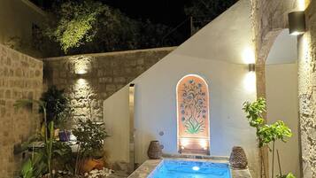 Une cour pavée de pierres, avec une piscine, des chaises longues en bois et des plantes en pot.