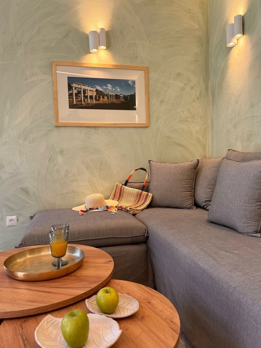 Ein gemütliches Wohnzimmer mit einem grauen Sofa, einem gerahmten Bild an der Wand und einem kleinen Holztisch mit einer Tablett und Äpfeln.