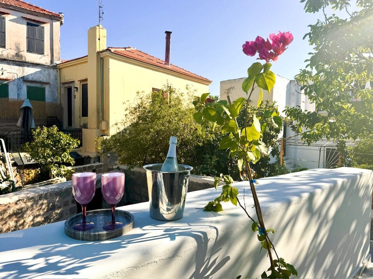 A table with two purple glasses, a metal container, and a pink flower on a sunlit terrace with buildings in the background.