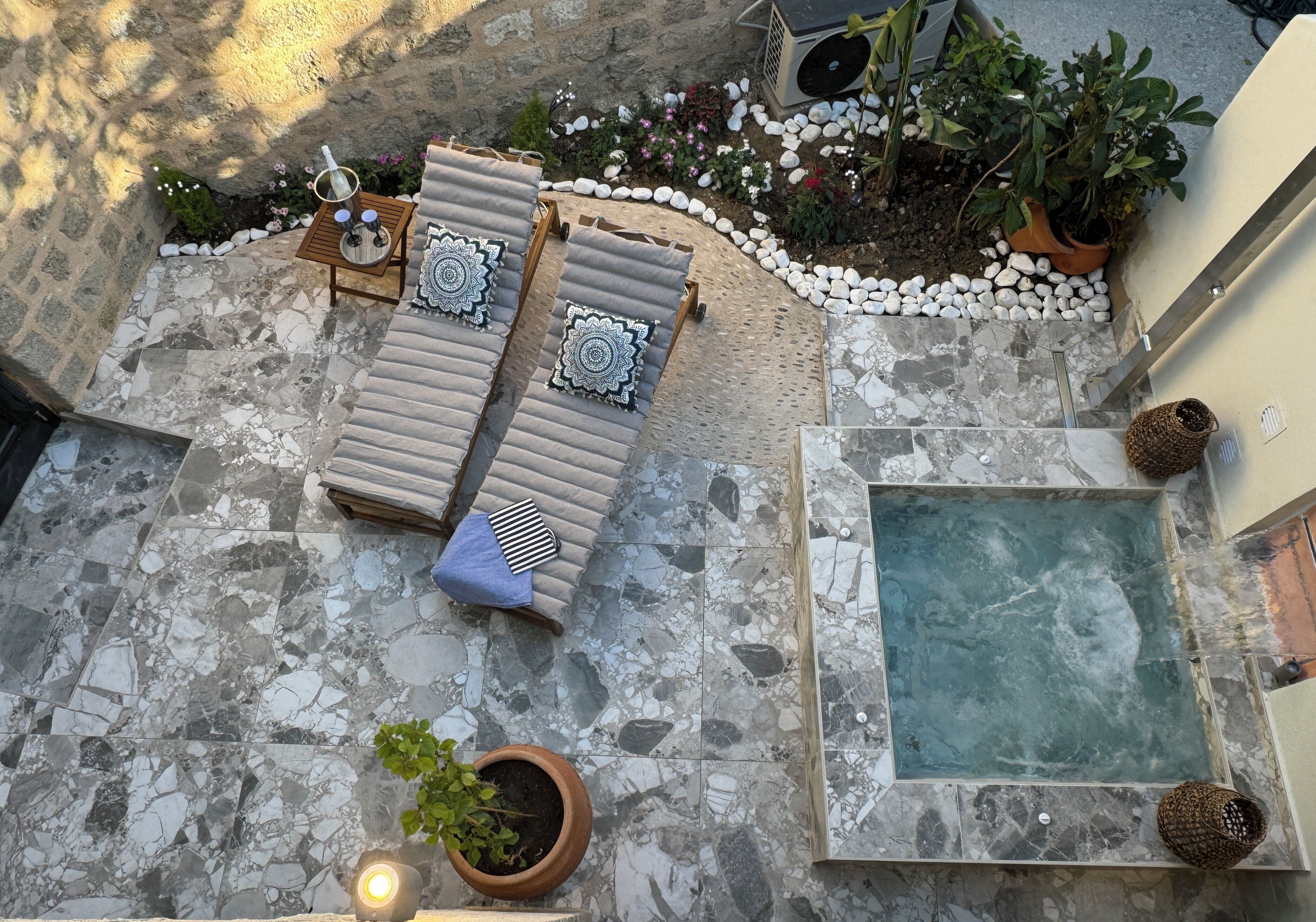Eine Dachterrasse mit Steinboden, zwei Liegestühle mit Kissen, einem kleinen Tisch und einer Flasche sowie einem Wasserspiel.