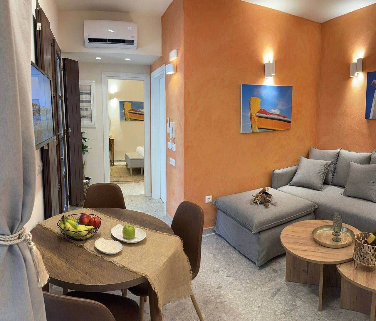 A modern living room with a grey sofa, a wooden dining table with a fruit bowl, and a small round table with a decorative item.