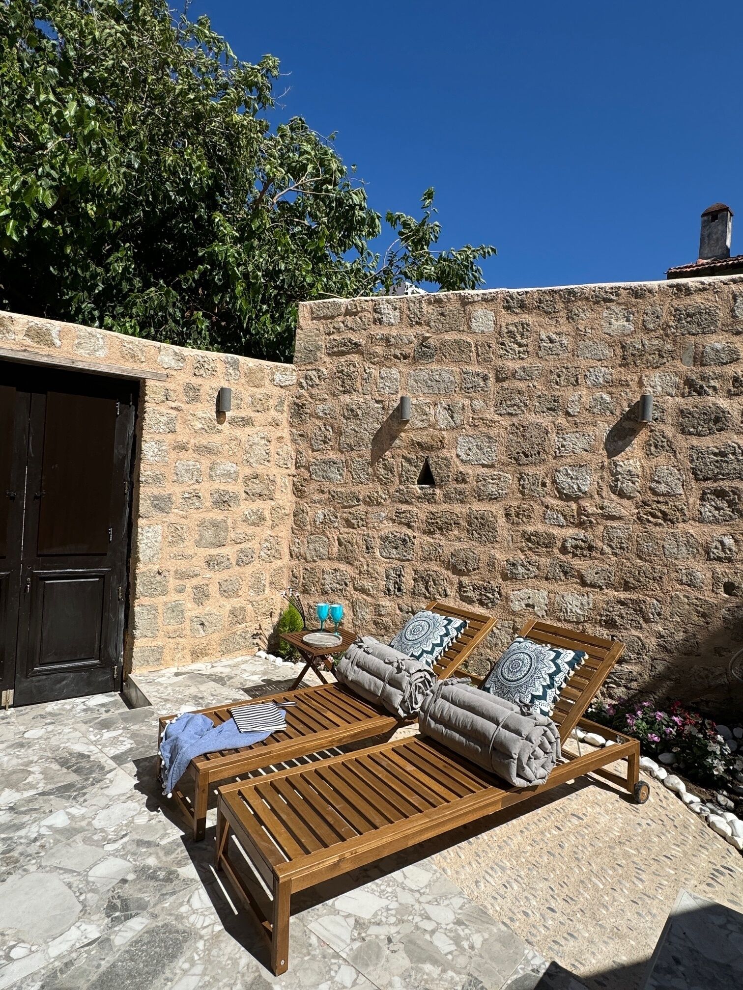 A stone wall with a wooden bench and cushions, a door, and a tiled patio.