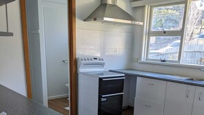 Una cocina con estufa blanca, horno y campana extractora de acero inoxidable. Hay una ventana con vistas al exterior, y el suelo es de madera.