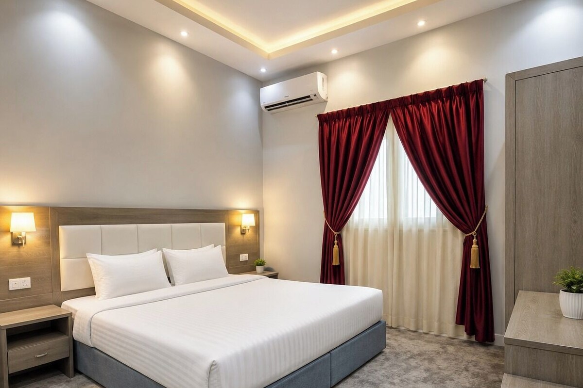 A hotel room with a large bed, bedside tables, an air conditioning unit, a desk with a chair, and a window with curtains.