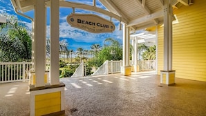Een overdekt buitengebied met een bord waarop 'Beach Club' staat, en palmbomen op de achtergrond.