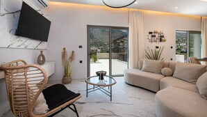 A modern living room with a sofa, a wicker chair, a glass coffee table, and a flat-screen TV mounted on the wall. - Esotico Escape Villa (Kournas)