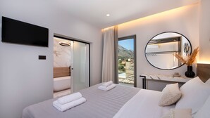 A modern hotel room with a large bed, a flat-screen TV, a bathroom with a glass door, a view of the mountains, and a round mirror on the wall. - Esotico Escape Villa (Kournas)