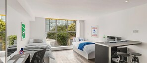 Un dormitorio moderno con cama, escritorio y silla. Amplias ventanas ofrecen vistas a zonas verdes.