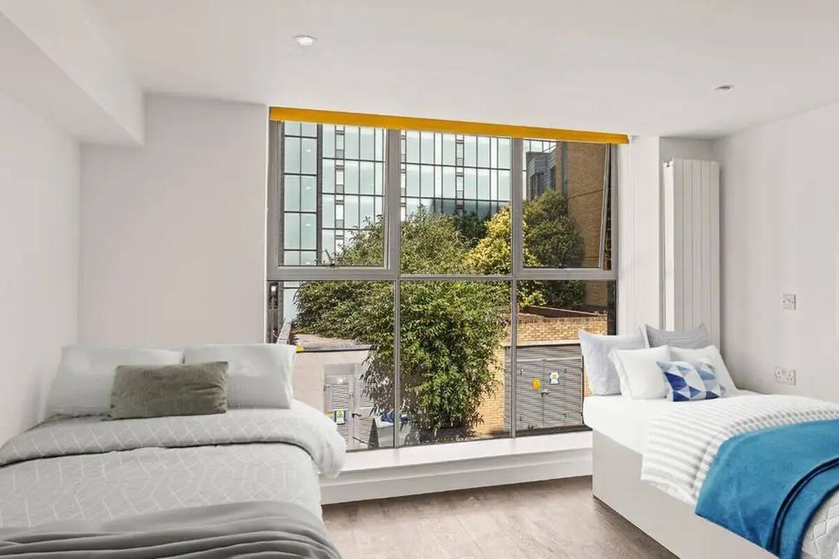 A modern hotel room with two beds, a large window with a view of the city, and a poster on the wall.