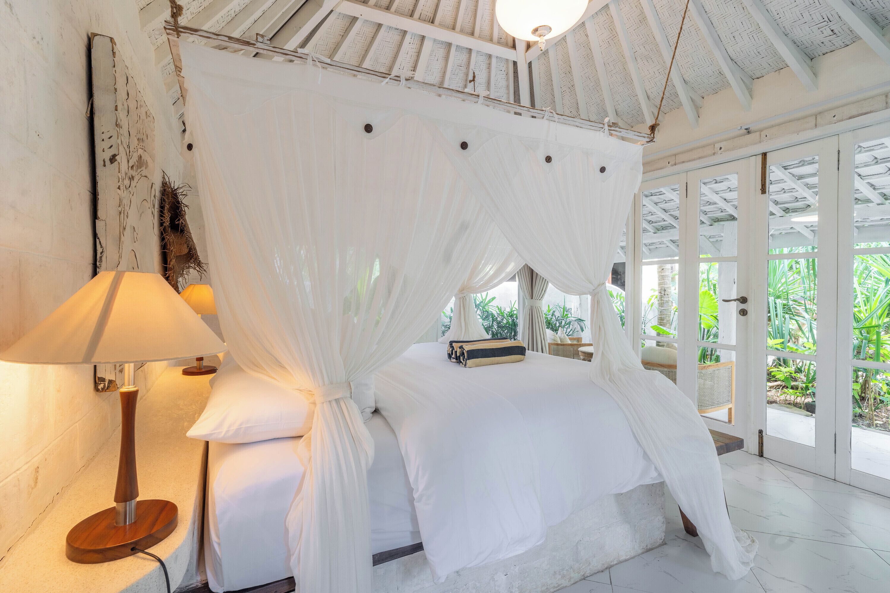 Un dormitorio con cama con dosel, una lámpara de noche y vistas a la vegetación por la puerta.