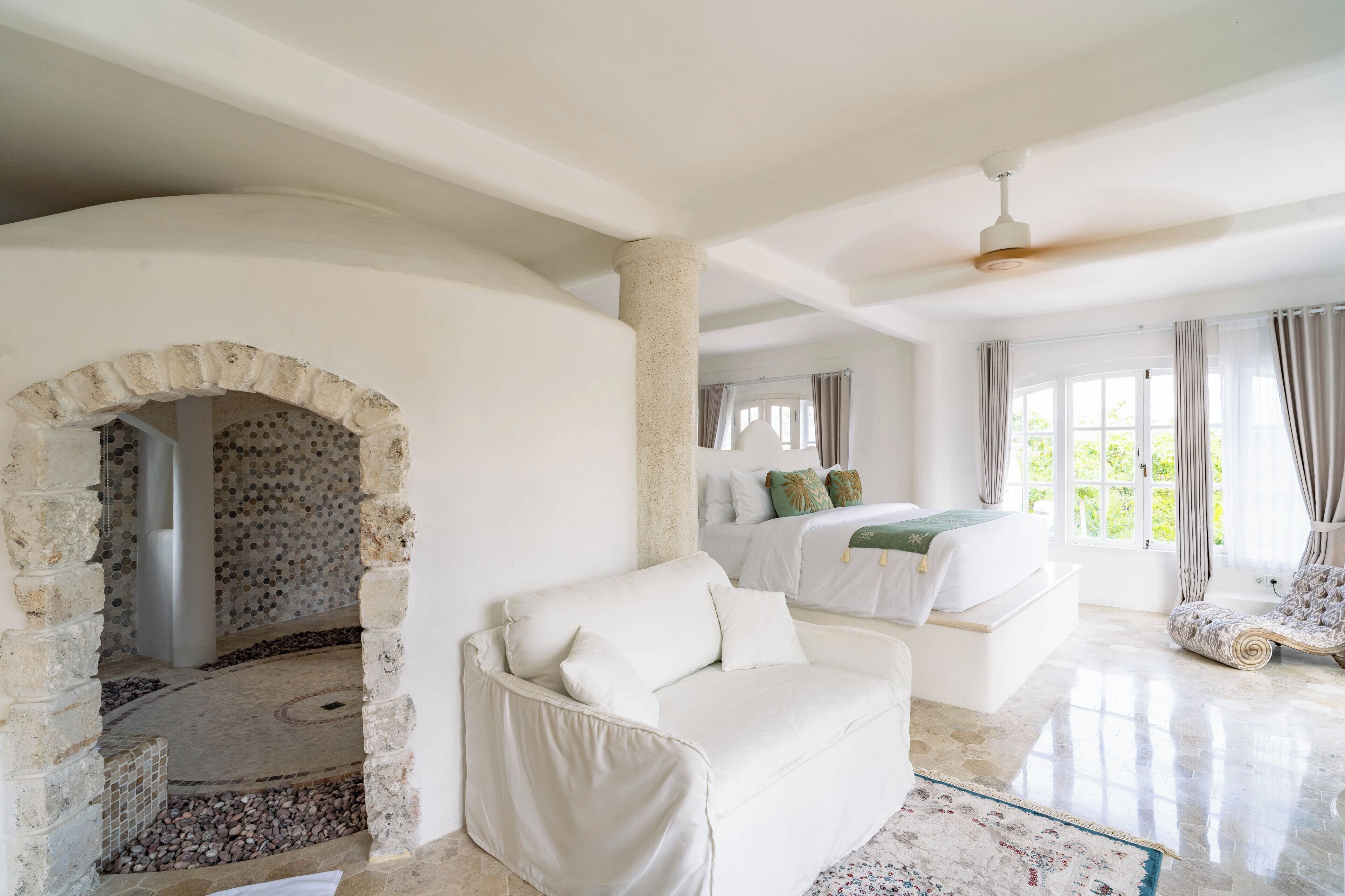A spacious room with a white sofa, a bed with green pillows, and a view of greenery outside.