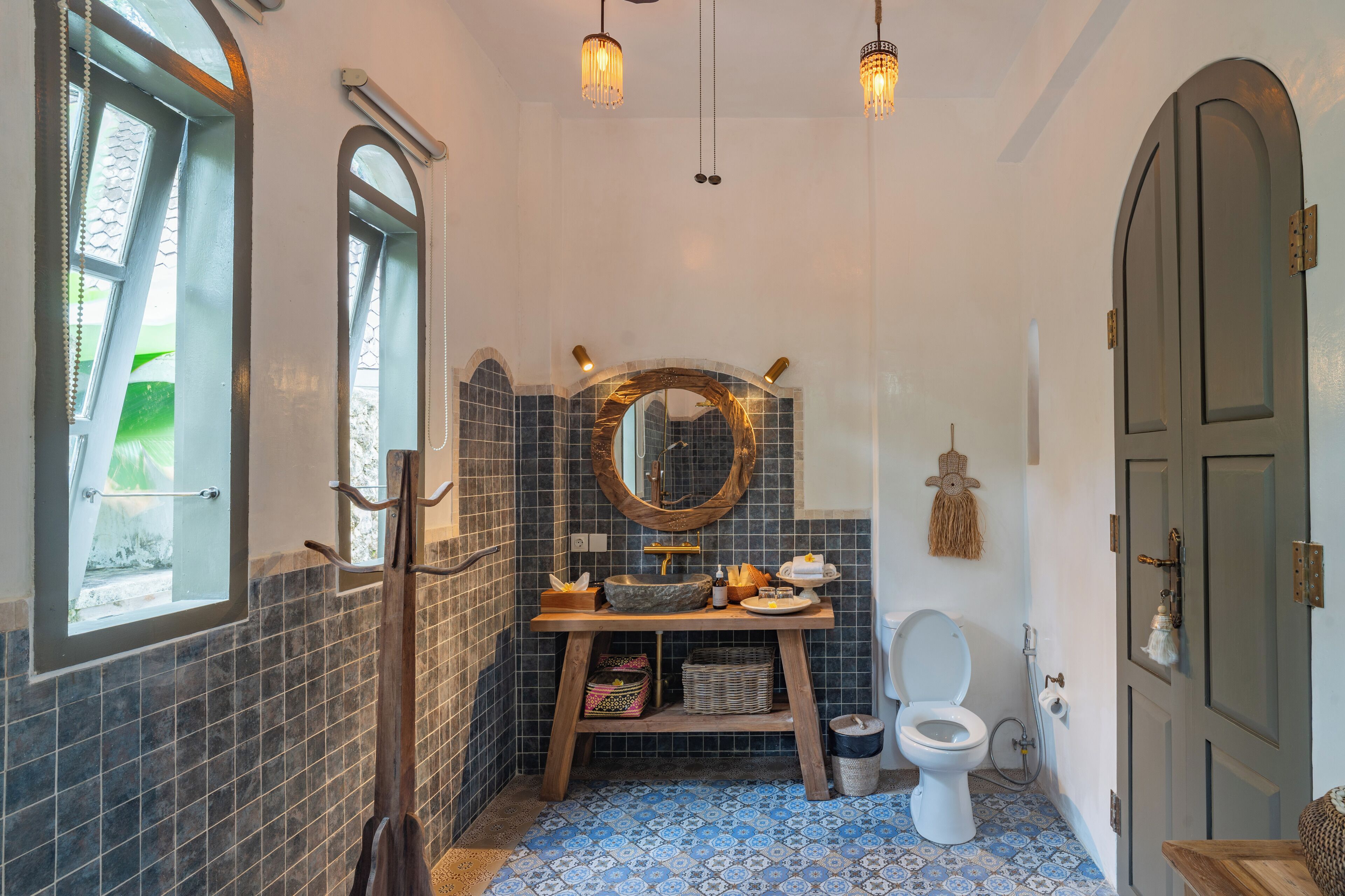 A bathroom with a wooden vanity, a round mirror, a white toilet, and a blue patterned floor.