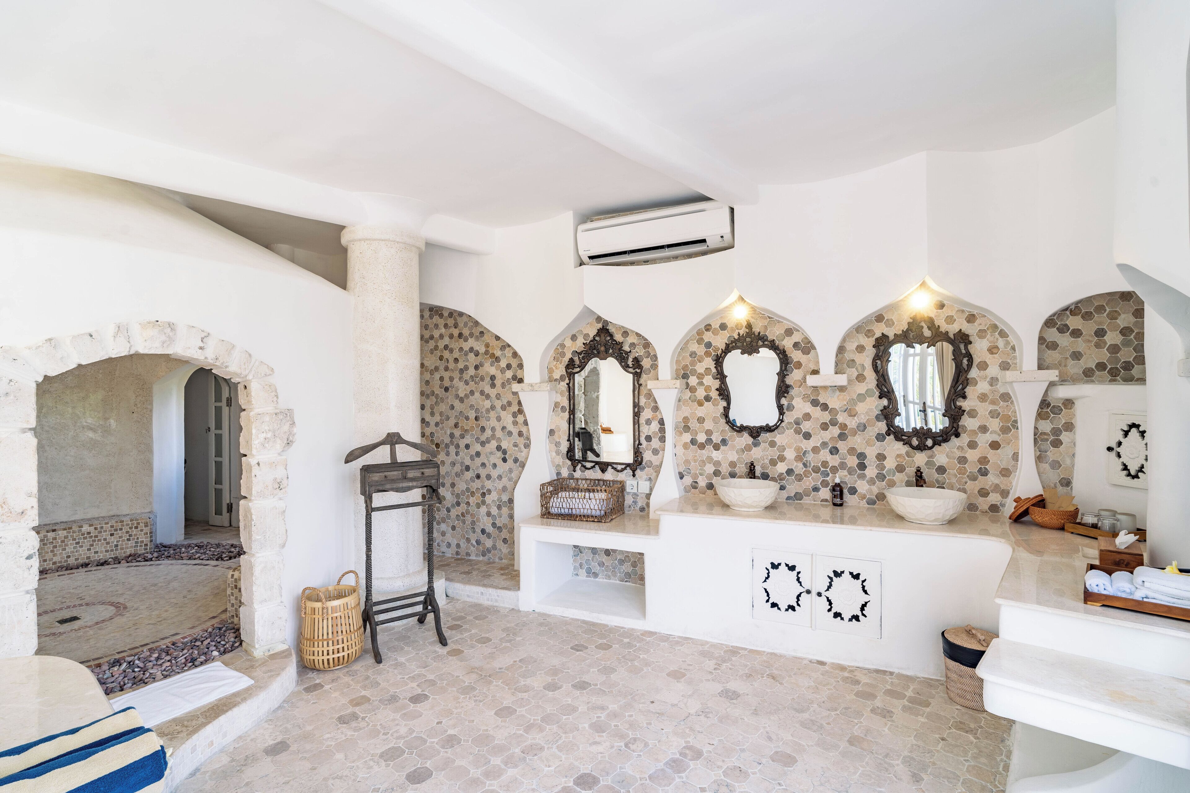 Un baño con suelo de piedra, paredes blancas y una zona central con tres espejos.