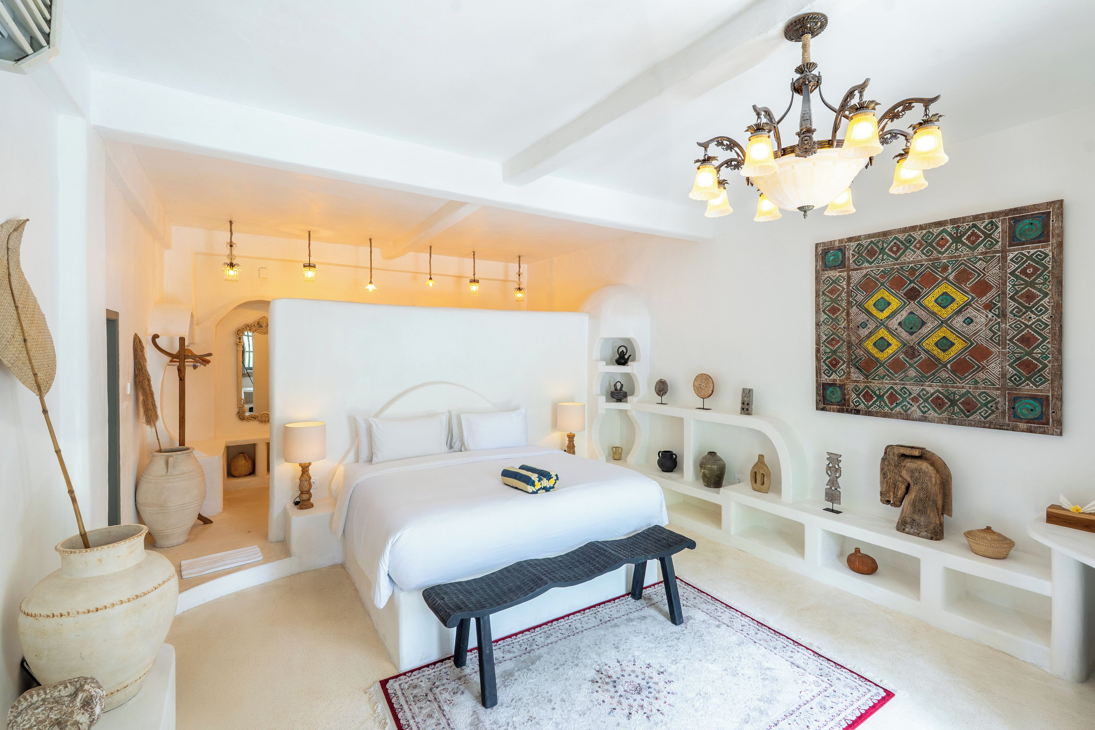 Een ruime slaapkamer met een groot bed, een bankje, een kroonluchter en een decoratief wandobject.