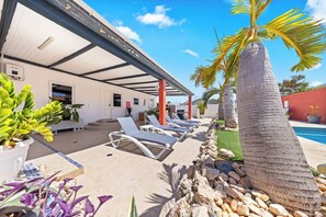 A covered outdoor area with lounge chairs, a pool, and palm trees. - Oranjestad Retreat: 4BR Paradise with Private Pool (Oranjestad)