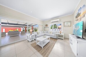 A spacious living room with a large window, a white sofa set, a coffee table, and a flat-screen TV.