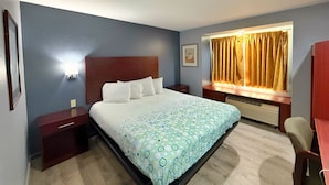 A hotel room with a large bed, a nightstand, a desk, a chair, a window with curtains, and a wall-mounted light.