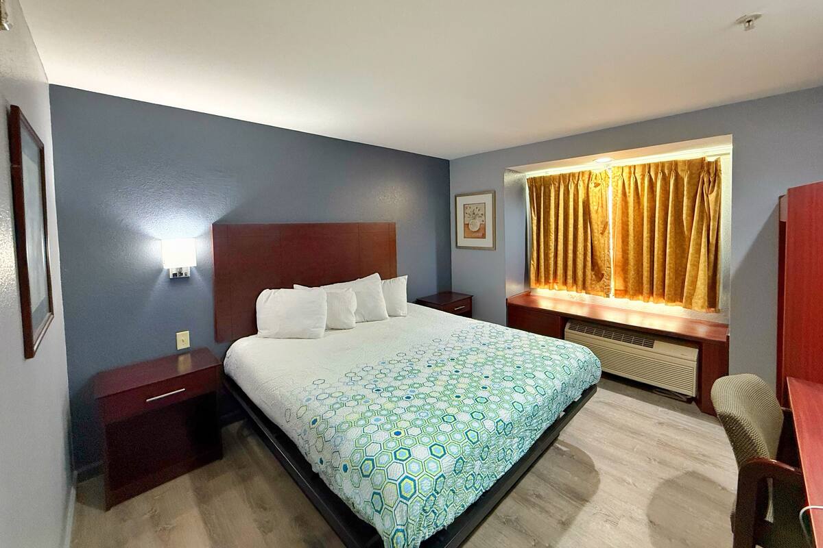 Habitación de hotel con una cama grande, una mesita de noche, un escritorio, una silla, un ventanal con cortinas y una lámpara de pared.