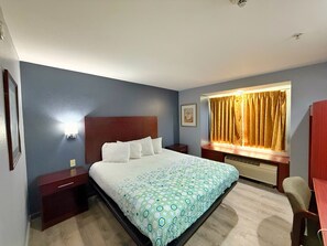 A hotel room with a large bed, a nightstand, a desk, a chair, a window with curtains, and a wall-mounted light.
