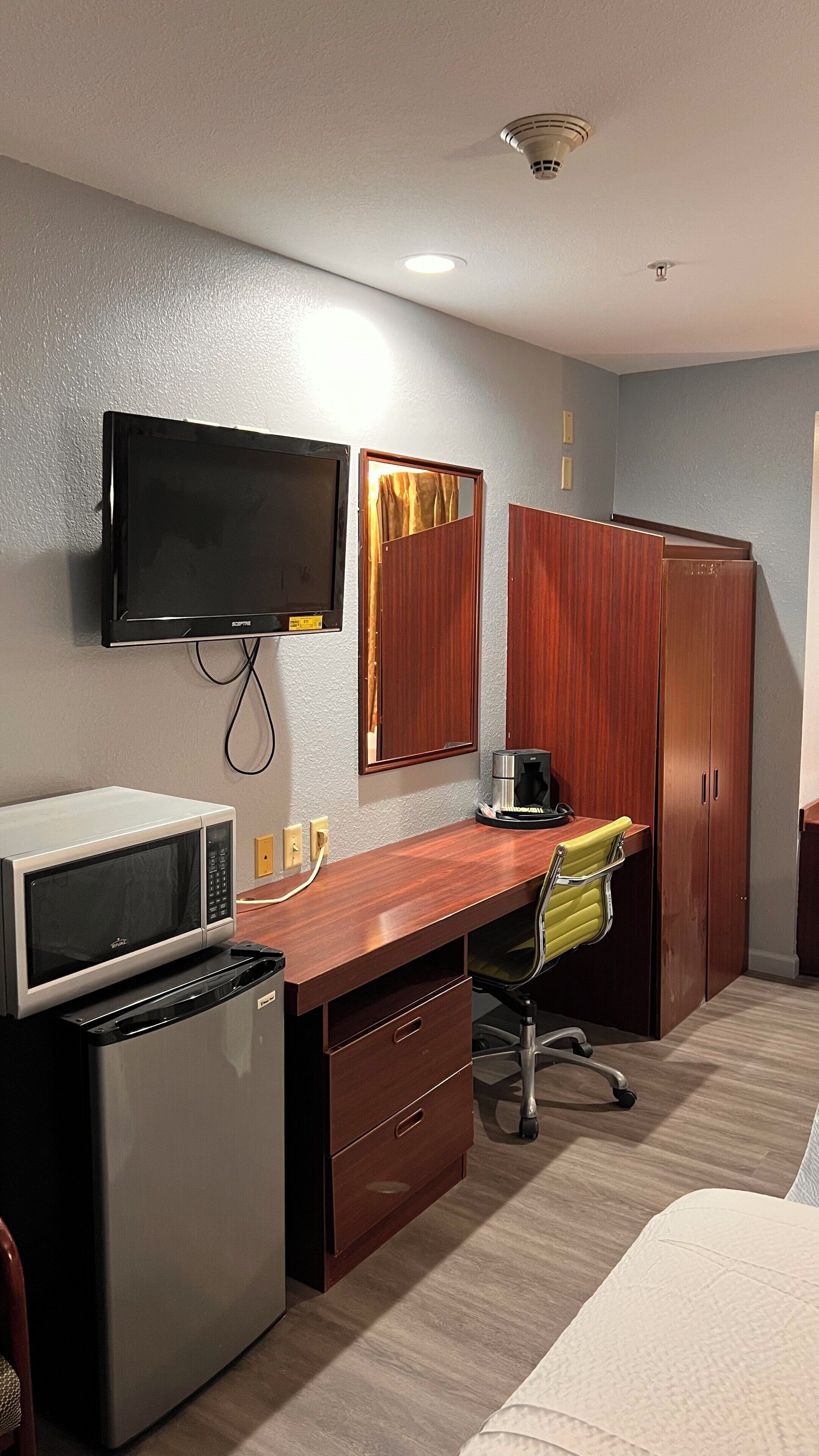 A hotel room with a wooden desk, a television mounted on the wall, a microwave, a refrigerator, a coffee maker, and a wardrobe.
