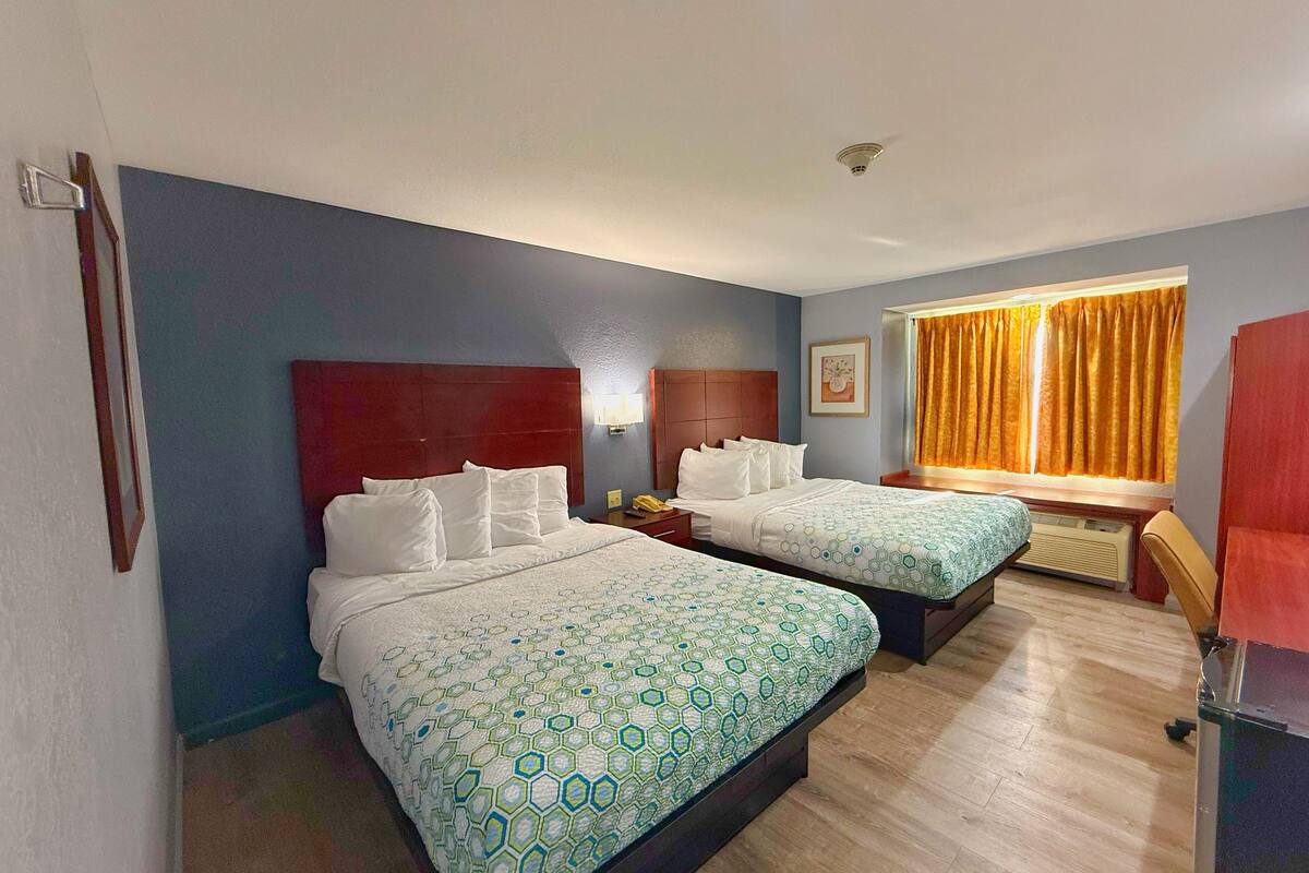 Habitación de hotel con dos camas, cabecero de madera, ventana con cortinas y mesita de noche con lámpara.